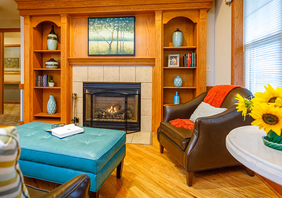 Cozy living room area with a lit fireplace surrounded by wooden built-in shelves holding decorative vases, books, and a framed photo. A brown leather armchair with a red throw blanket and a gray pillow sits next to a round white table with a vase of sunflowers. In front of the fireplace is a large turquoise ottoman with an open book and glasses on top. A painting of trees hangs above the fireplace.