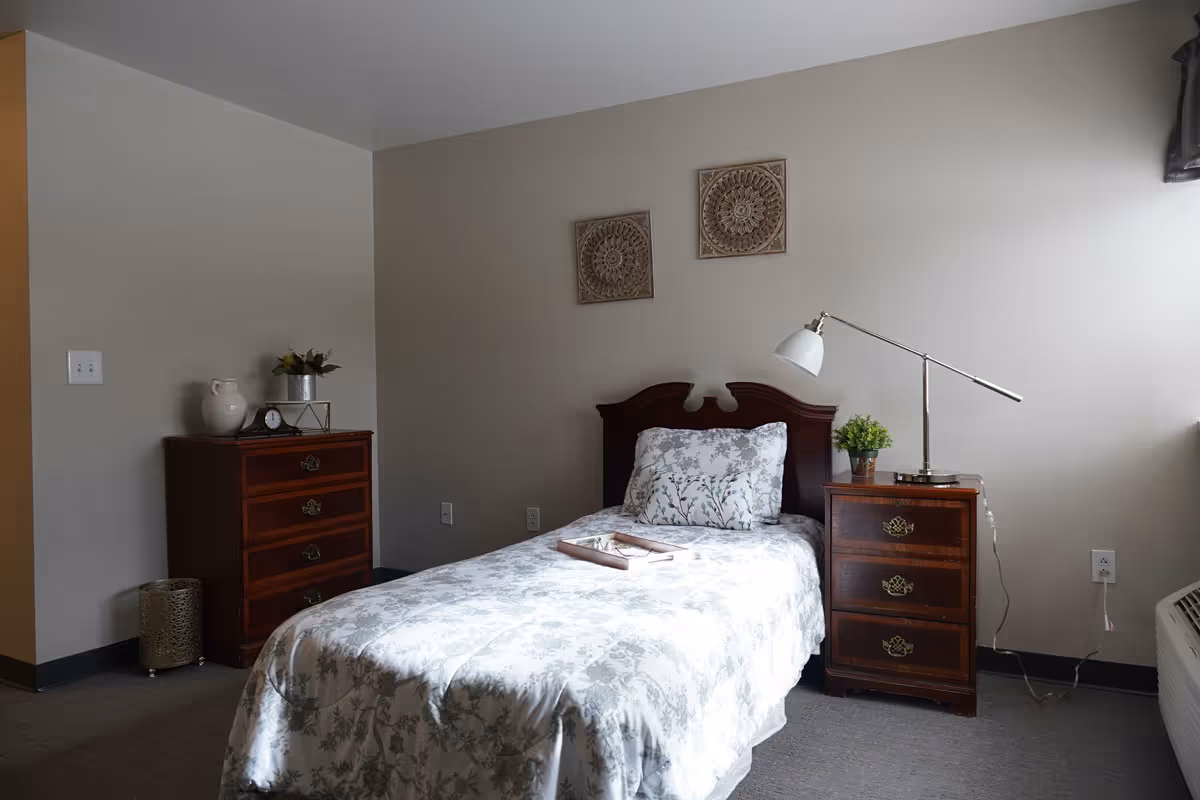 A neatly made single bed with floral patterned bedding and a matching pillow in a simple bedroom. The bed has a dark wooden headboard and is flanked by two wooden nightstands, one with a modern white desk lamp and a small green plant. On the opposite side, there is a wooden dresser with decorative items including a white pitcher, a clock, and a small plant. Two decorative wall hangings are above the bed on a plain light-colored wall.