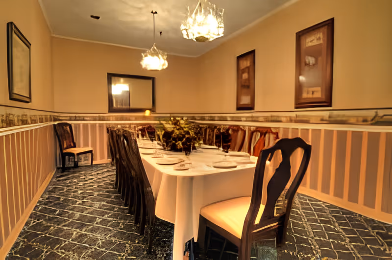 A formal dining room with a long rectangular table covered with a white tablecloth, set with plates, glasses, and napkins. The room has patterned dark flooring, beige walls with a decorative border, and framed artwork. There are multiple wooden chairs with cushioned seats around the table, and two hanging light fixtures above.