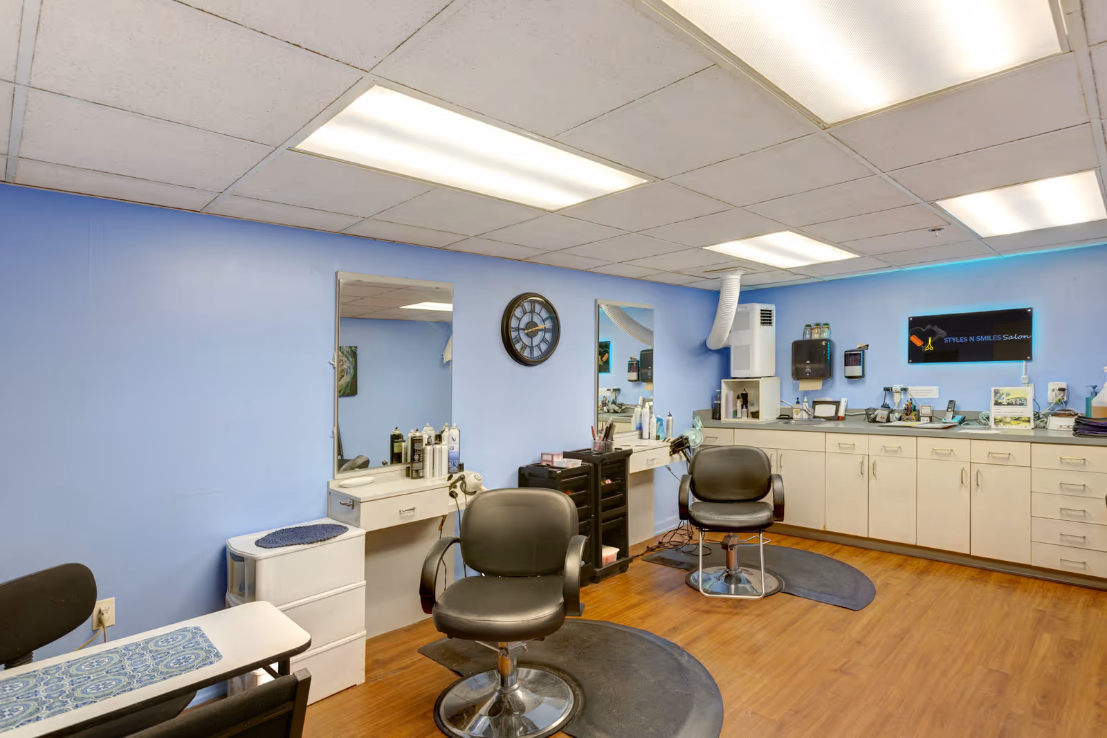Interior of a salon room with two black salon chairs in front of mirrors mounted on a light blue wall. The room has wooden flooring, white cabinets along one wall, and various hair care products and equipment on the counters. A clock is mounted on the wall between the mirrors, and fluorescent ceiling lights illuminate the space.