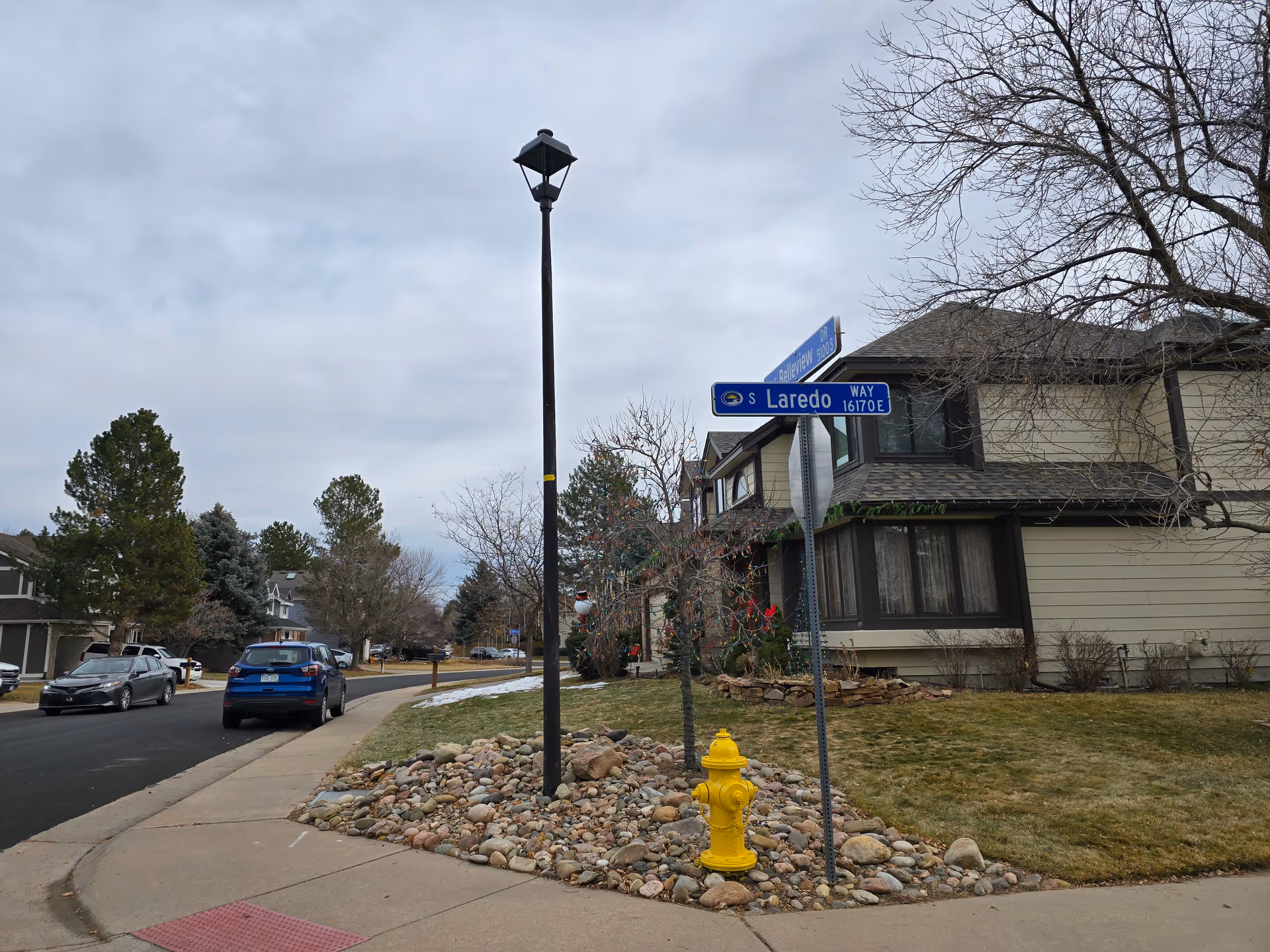 A suburban street corner with a yellow fire hydrant, a street lamp, and street signs for S Laredo Way and another street. There are houses with trees and parked cars along the road under a cloudy sky.