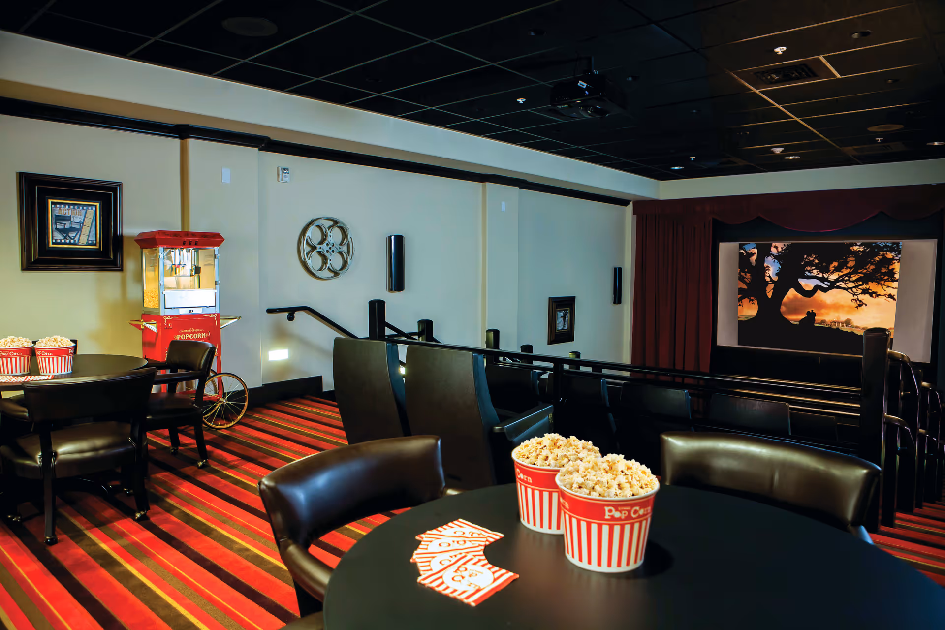 Interior of a small movie theater room with a screen showing a silhouette of a tree and a person. The room has tiered seating with black chairs, a red and black striped carpet, a popcorn machine, and tables with buckets of popcorn and movie tickets.