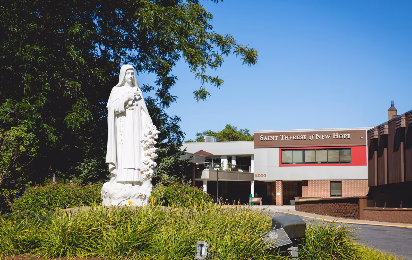 Outdoor view of Saint Therese Senior Living of New Hope building with a large white statue of a saint holding flowers in the foreground surrounded by greenery and trees under a clear blue sky.