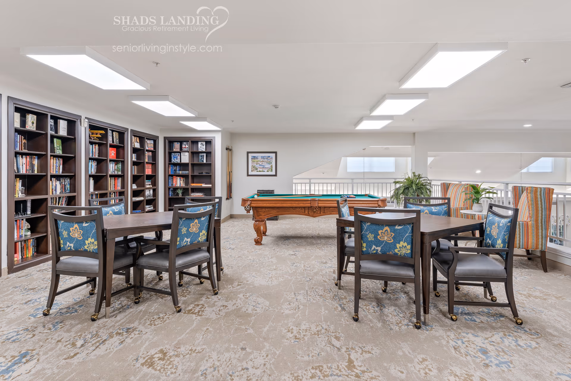 A bright and spacious common area in a retirement living facility featuring two tables with four chairs each, a pool table in the center, bookshelves filled with books along the left wall, and two colorful armchairs with plants near the railing on the right side.