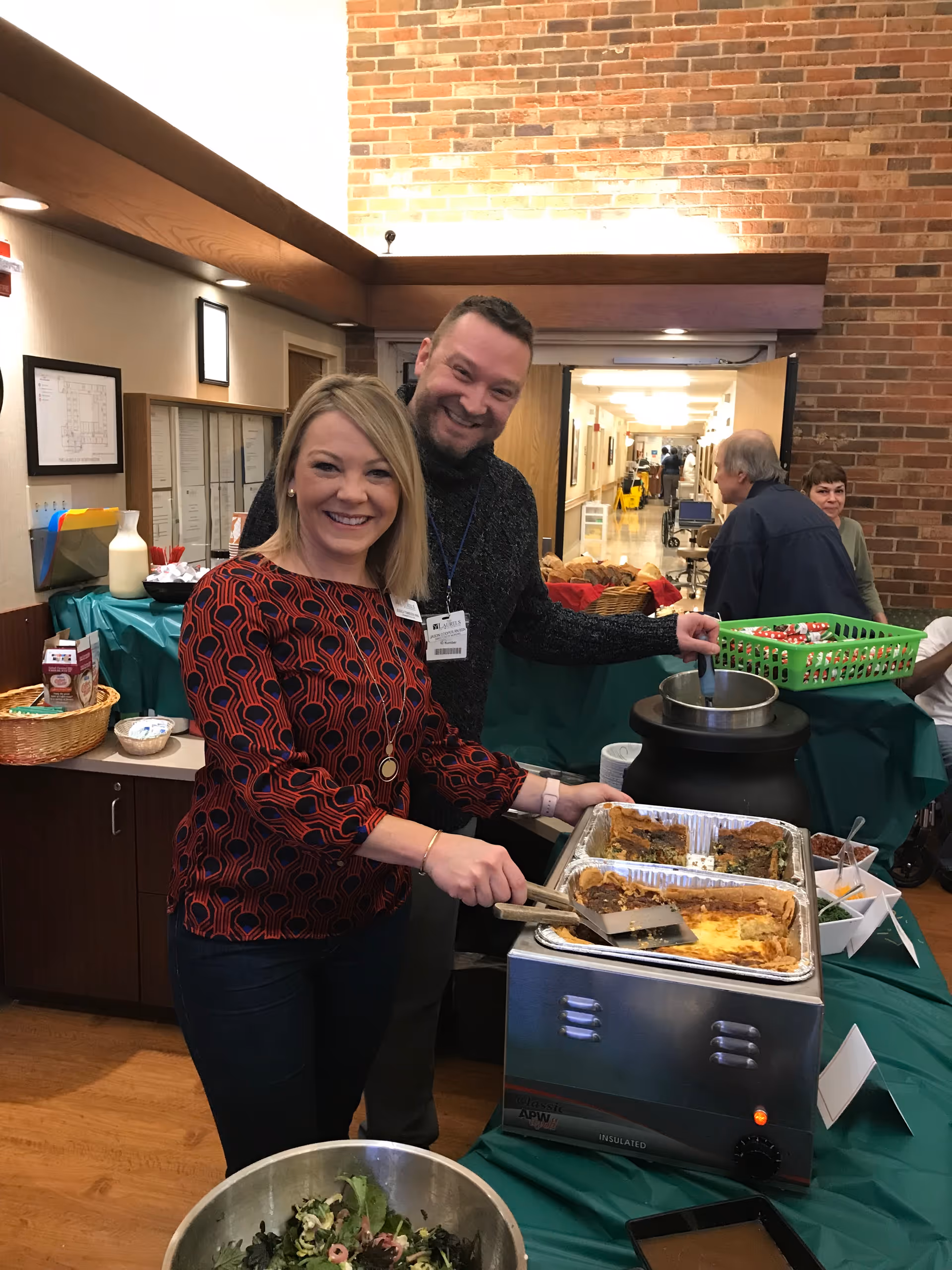 Two smiling adults, a woman and a man, serving food from trays on a buffet table covered with a green tablecloth in an indoor setting with brick walls. The woman is holding a spatula and serving quiche or similar food, while the man is holding a ladle near a soup pot. Other people and food items are visible in the background.