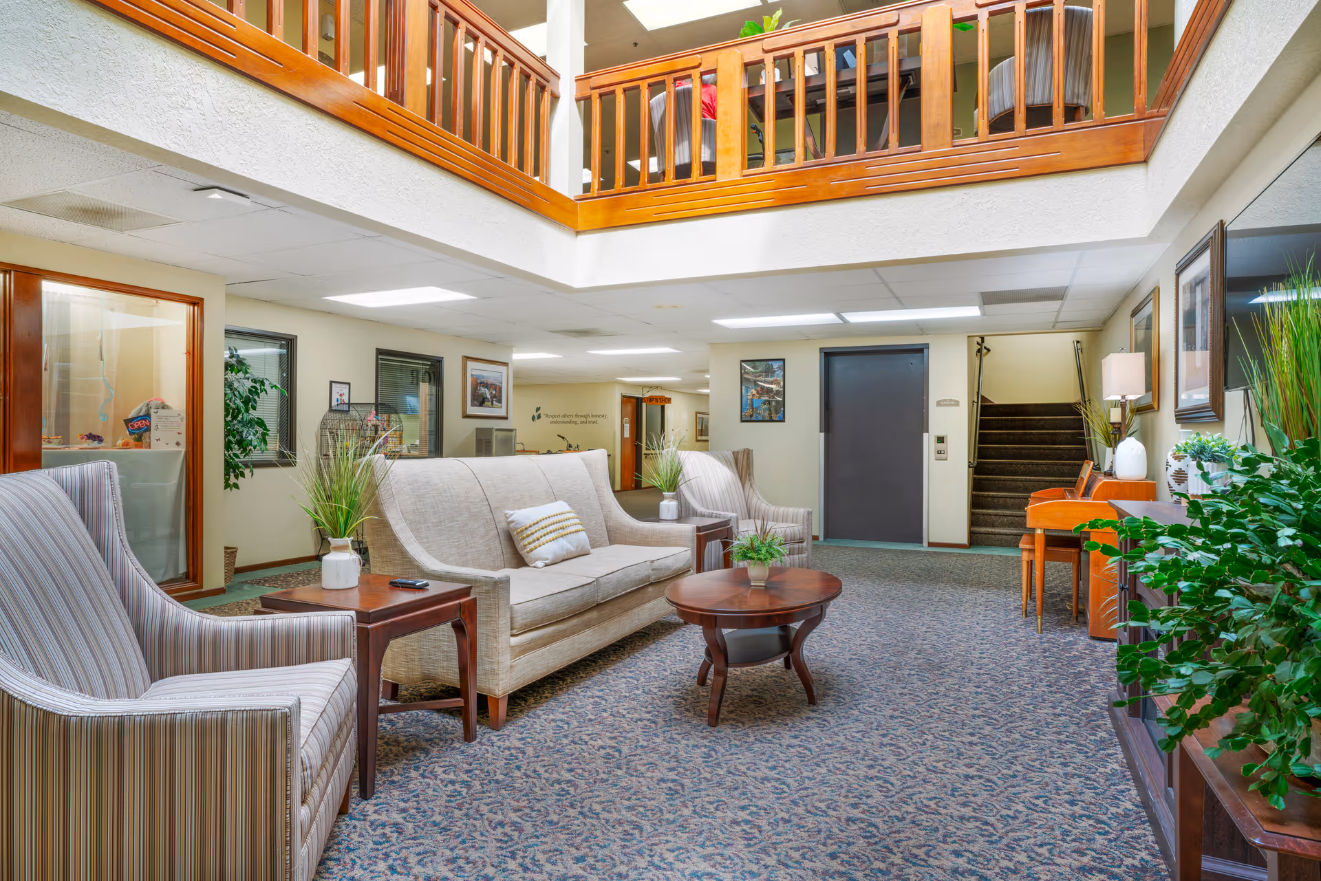A bright and spacious indoor common area with comfortable seating including a beige sofa and two striped armchairs around wooden tables. The room has carpeted flooring, potted plants, framed pictures on the walls, and a staircase leading upstairs. A wooden railing balcony overlooks the area from above.