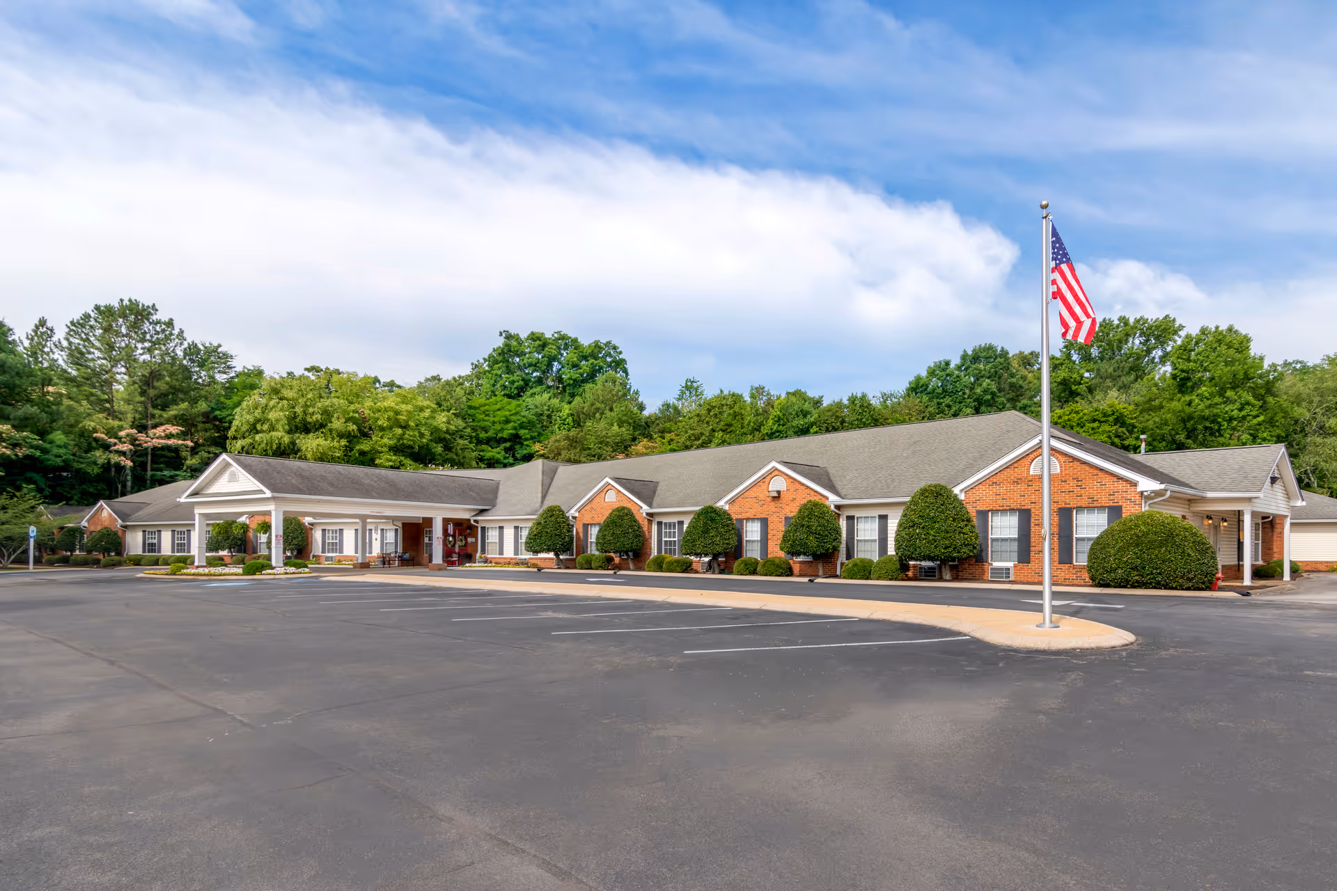 Exterior view of a single-story brick building with a covered entrance, neatly trimmed bushes, an American flag on a flagpole, and a large empty parking lot in front under a partly cloudy sky.
