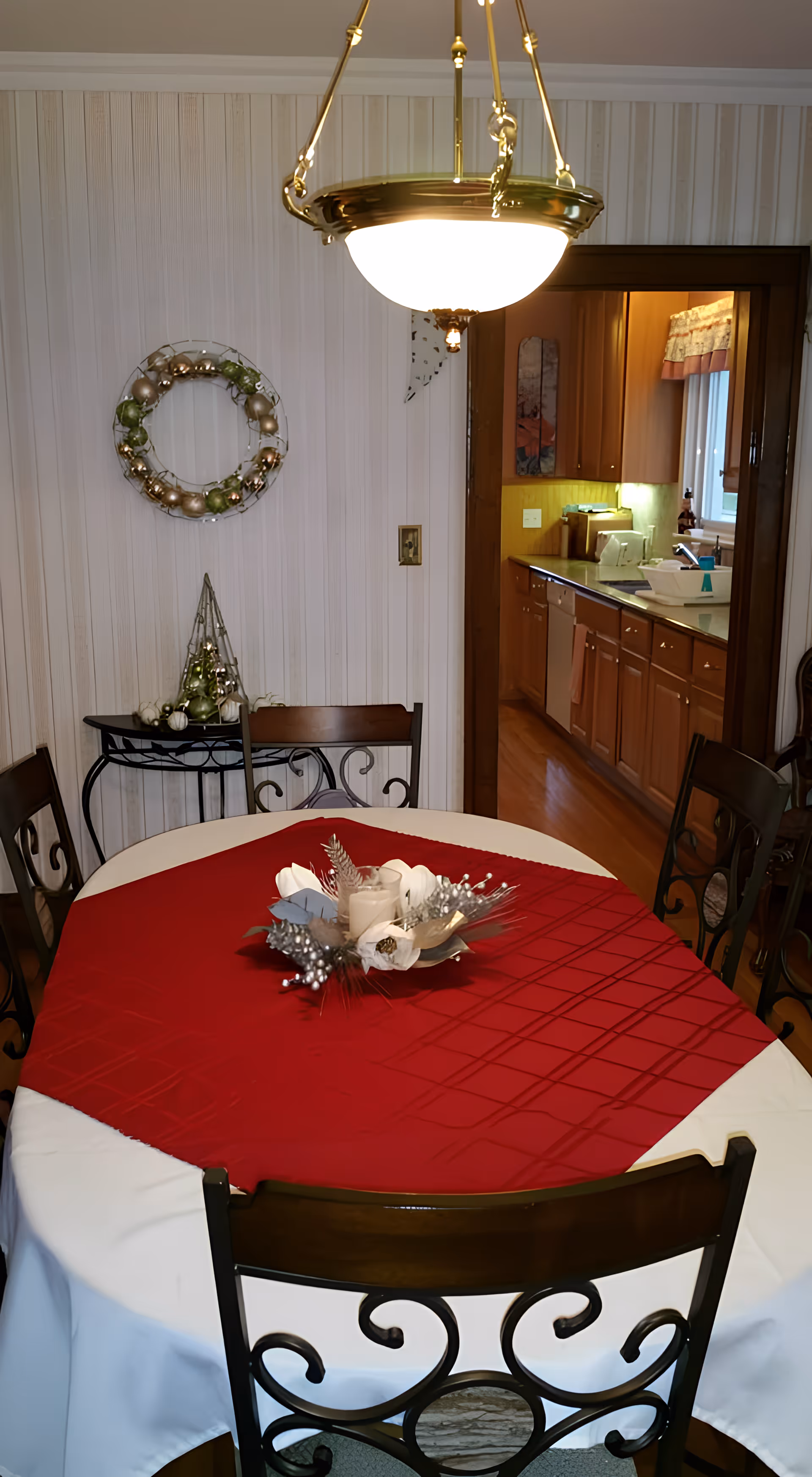 A dining room with a round table covered with a white tablecloth and a red table runner. The table has a decorative centerpiece with candles and silver ornaments. Four chairs with ornate metal backs surround the table. A hanging light fixture is above the table. In the background, there is a doorway leading to a kitchen with wooden cabinets and a window with a valance. On the left wall, there is a small black table with holiday decorations and a wreath hanging above it.