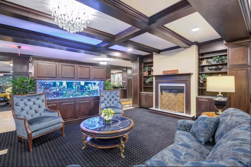 A cozy living room with dark wooden beams on the ceiling, a crystal chandelier, and a large built-in aquarium. The room features two blue upholstered armchairs, a blue patterned sofa, a round coffee table with a floral centerpiece, a fireplace with a decorative screen, built-in wooden shelves with plants and decor, and a table lamp.