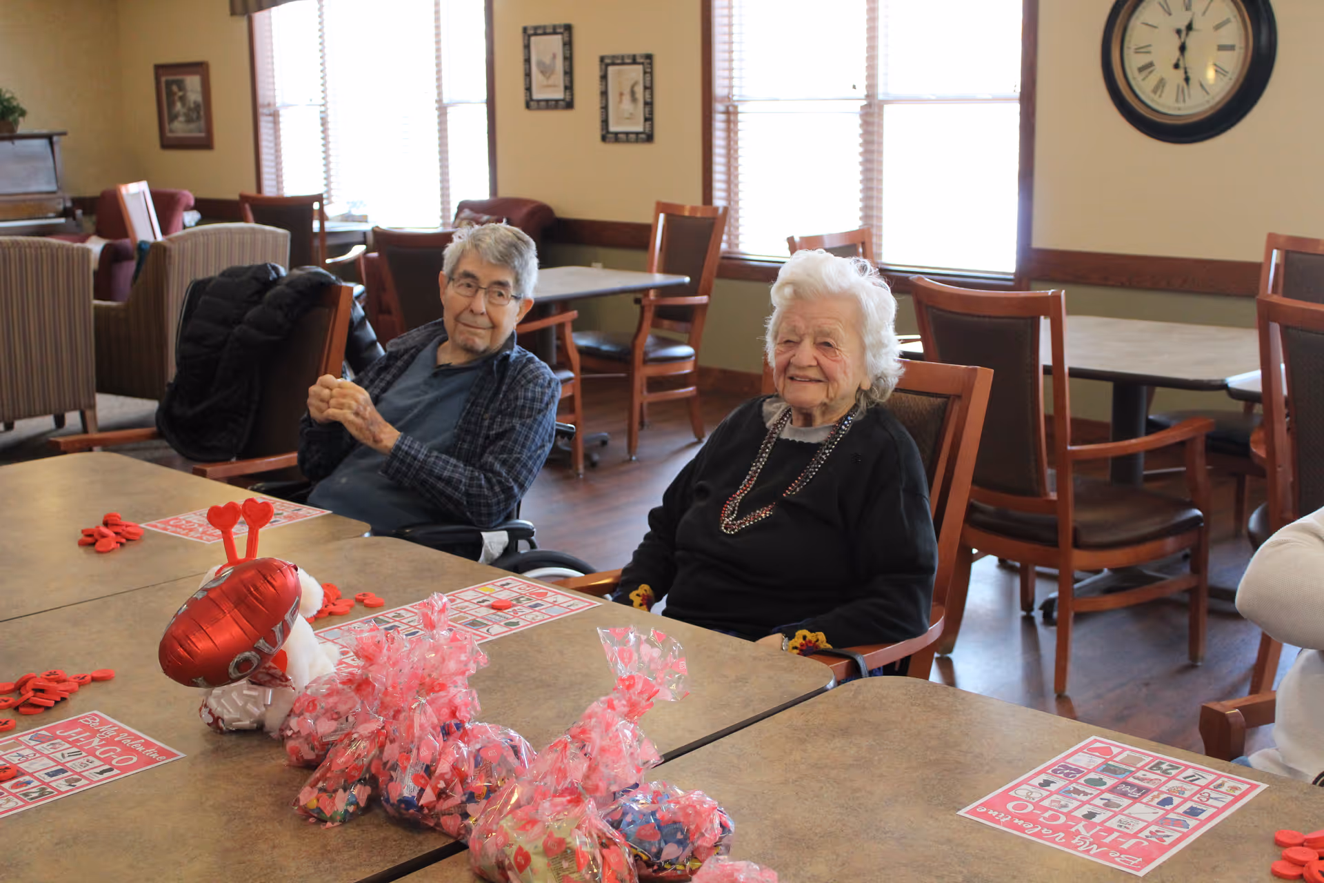 Two elderly residents sitting at a table in a communal activity room with bingo cards and Valentine's decorations.