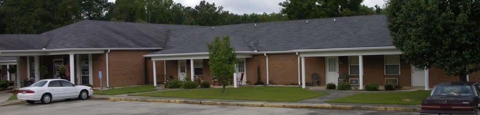 Exterior view of a single-story brick building with a dark shingled roof, several windows, and doors. There are two cars parked in front on a paved area, with small trees and grass patches along the building. The sky is overcast and trees are visible in the background.