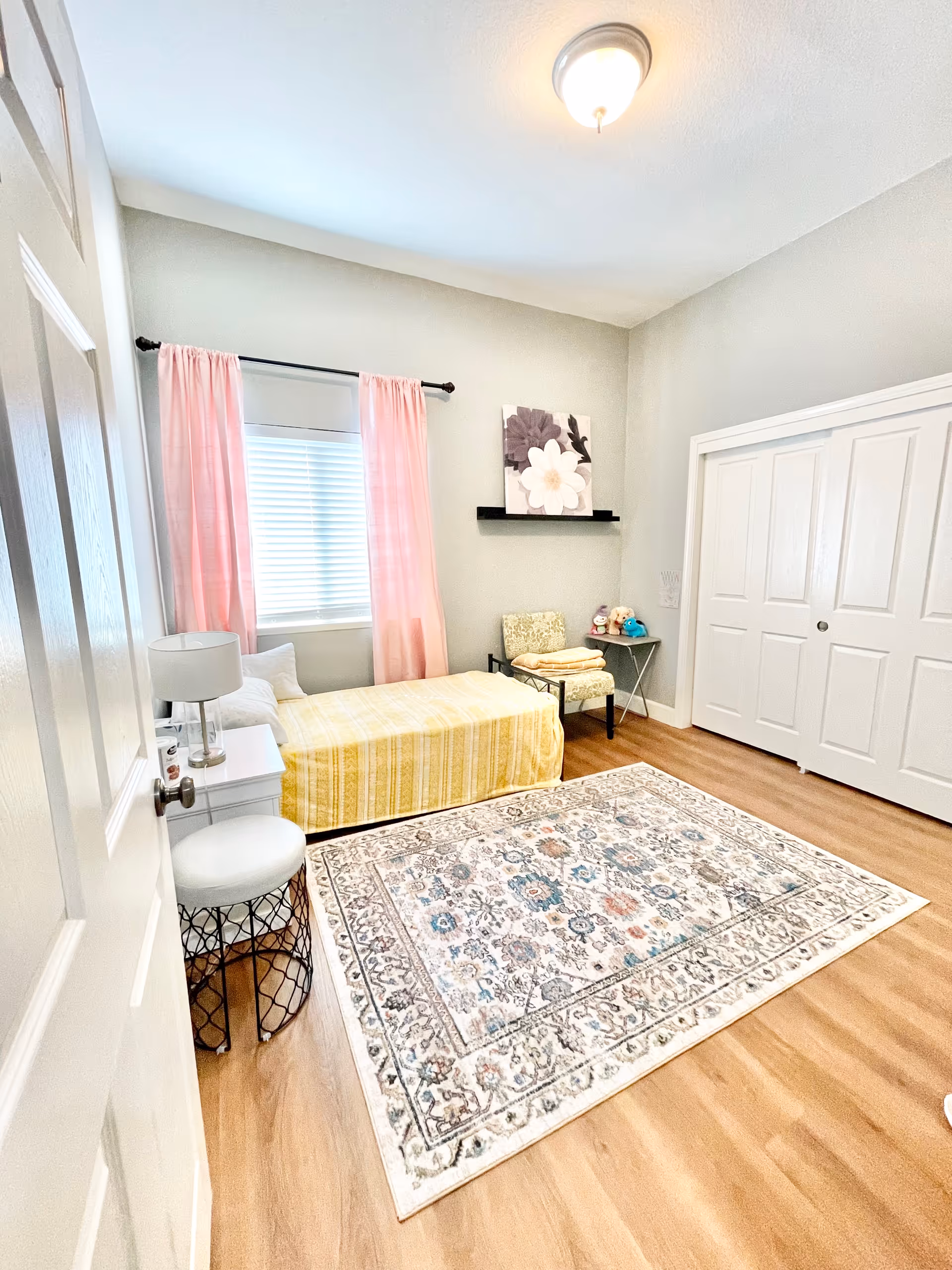 A bright and cozy bedroom with light gray walls, a single bed with a yellow striped bedspread, a white nightstand with a lamp, a small round stool, a patterned area rug on wooden flooring, pink curtains on the window, a floral wall art above a chair with folded towels, and a closet with white sliding doors.