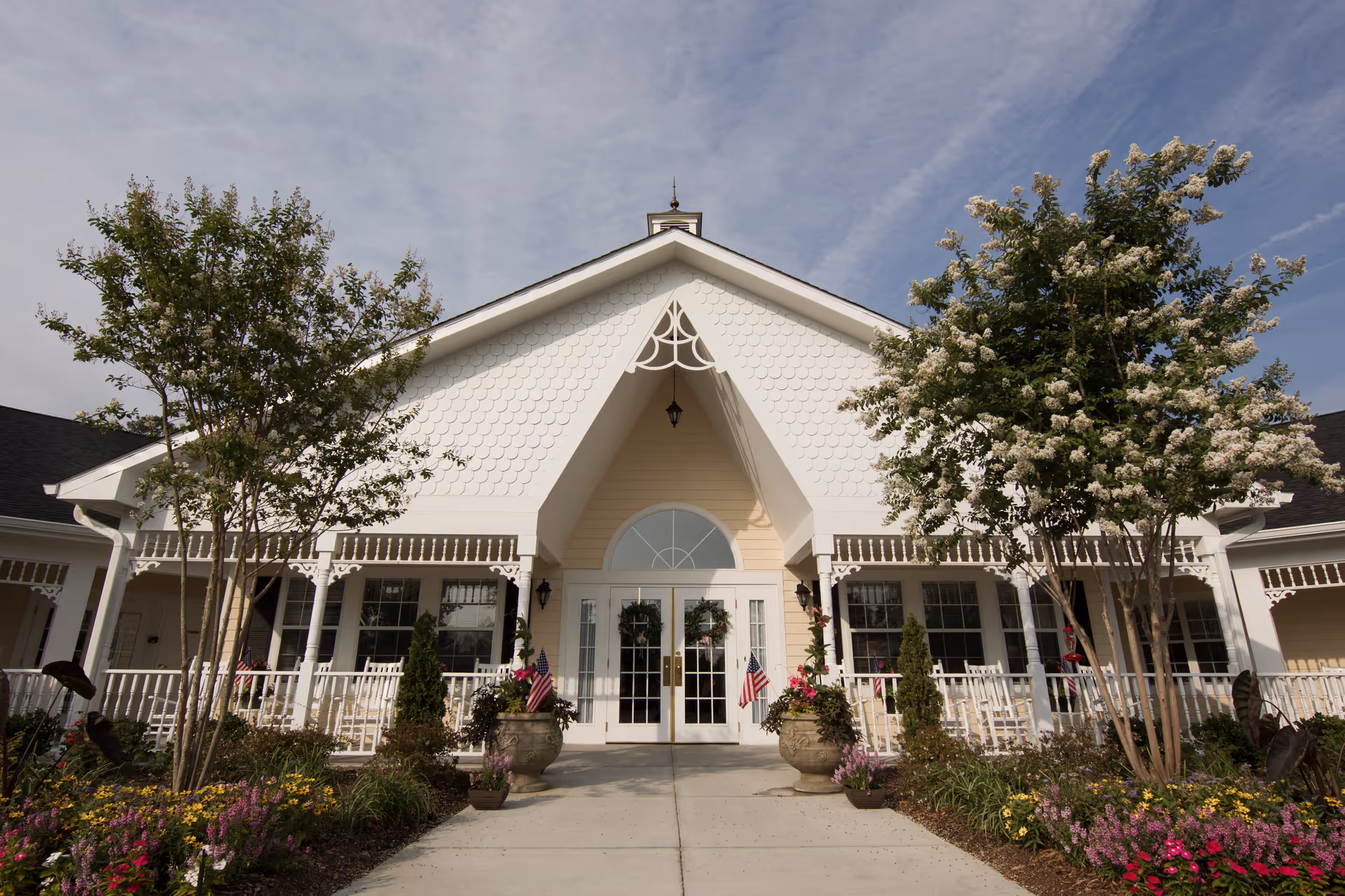 Front exterior view of a senior living facility with a white peaked roof, decorative trim, and a porch with white railings. The entrance has double glass doors adorned with wreaths and flanked by two large planters with American flags. There are flowering plants and trees on either side of the walkway leading to the entrance under a partly cloudy sky.