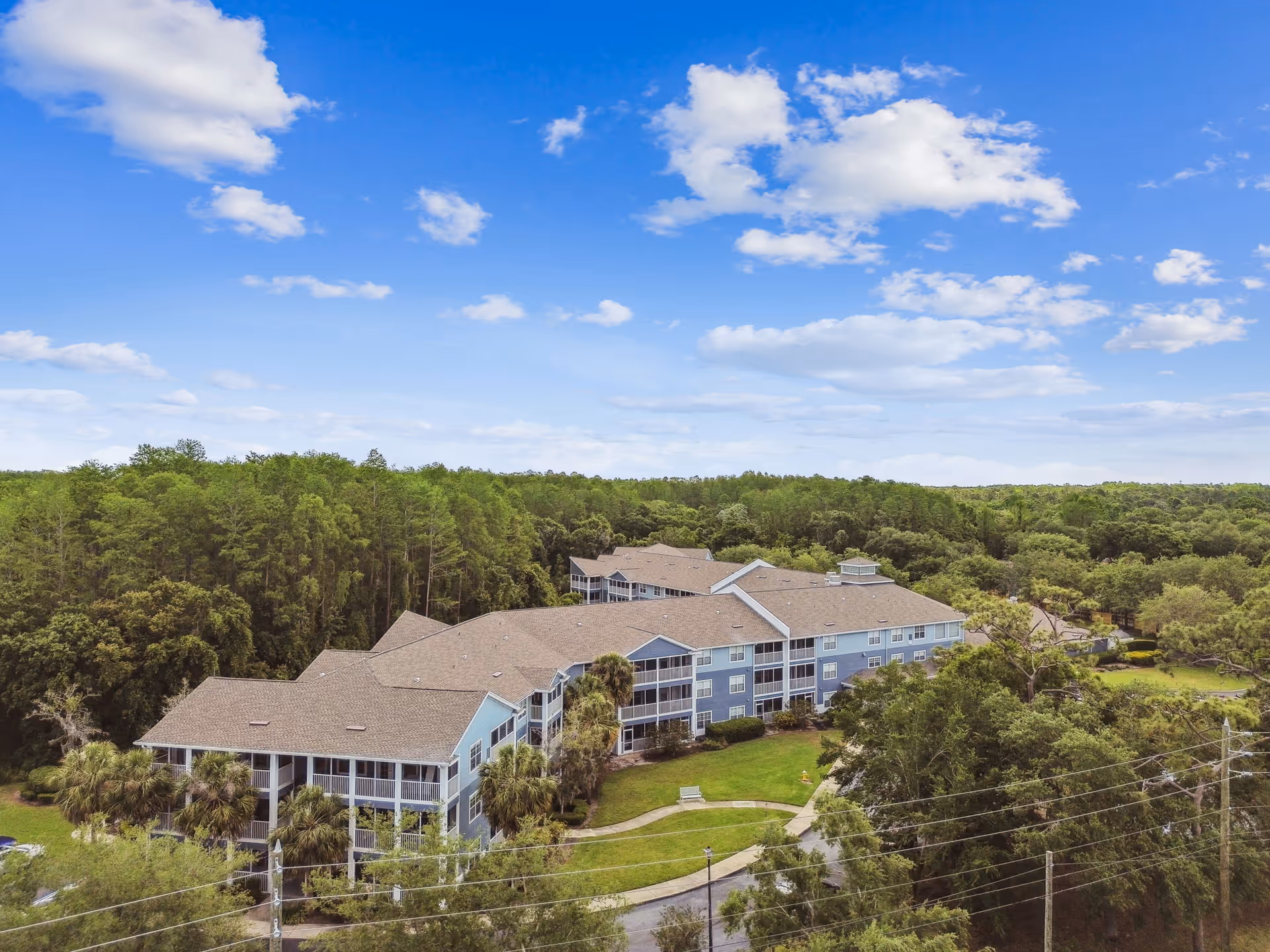 Aerial view of a large senior living facility building surrounded by lush green trees and landscaping under a blue sky with scattered clouds.