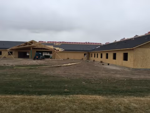 Under-construction, single-story facility with exposed framing and plywood sheathing beside a grassy field.