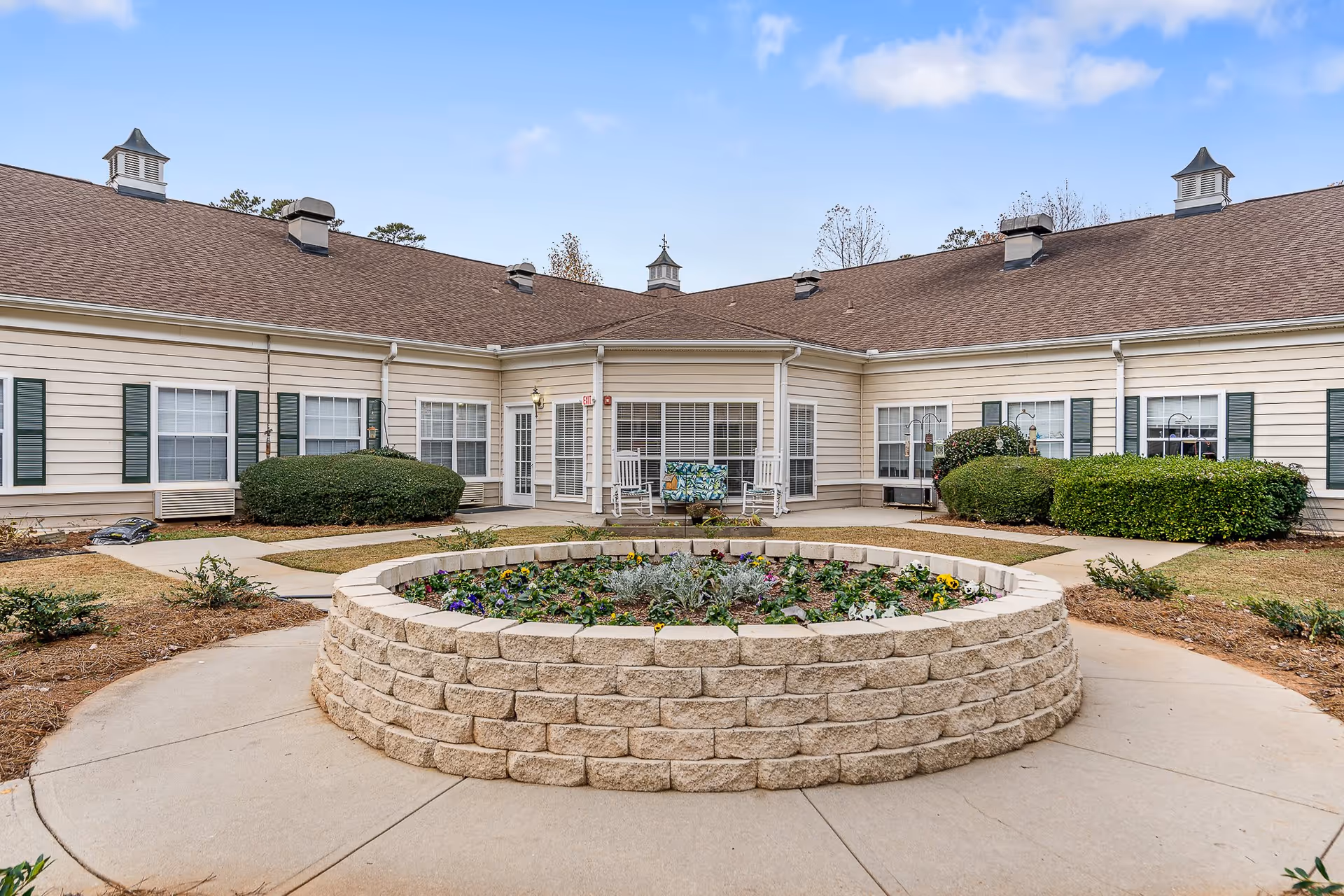 Outdoor courtyard area of a senior living facility with a circular raised flower bed in the center, surrounded by a concrete walkway. The building has beige siding, green shutters, and multiple windows. There are two white rocking chairs and a bench near the entrance doors.
