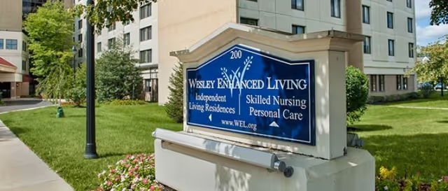 Sign reading 'Wesley Enhanced Living' in front of a multi-story senior living building with lawn and sidewalk.