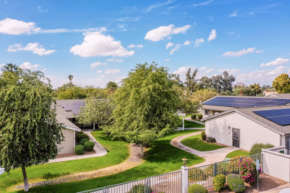View of a senior living community with single-story buildings featuring solar panels on the roofs, surrounded by green lawns, trees, and paved walkways under a partly cloudy blue sky.