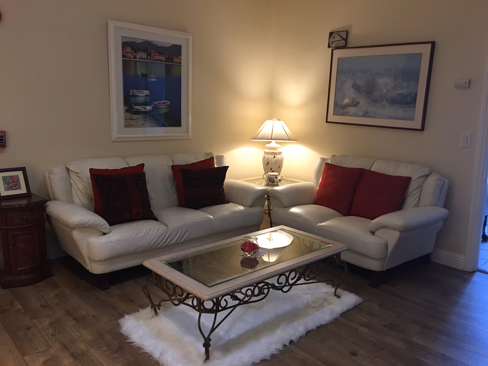 A cozy living room with two white leather sofas adorned with red and dark patterned cushions. Between the sofas is a glass-top coffee table with an ornate metal frame, placed on a white fluffy rug. A table lamp on a small corner table illuminates the space, and two framed paintings hang on the walls above the sofas.