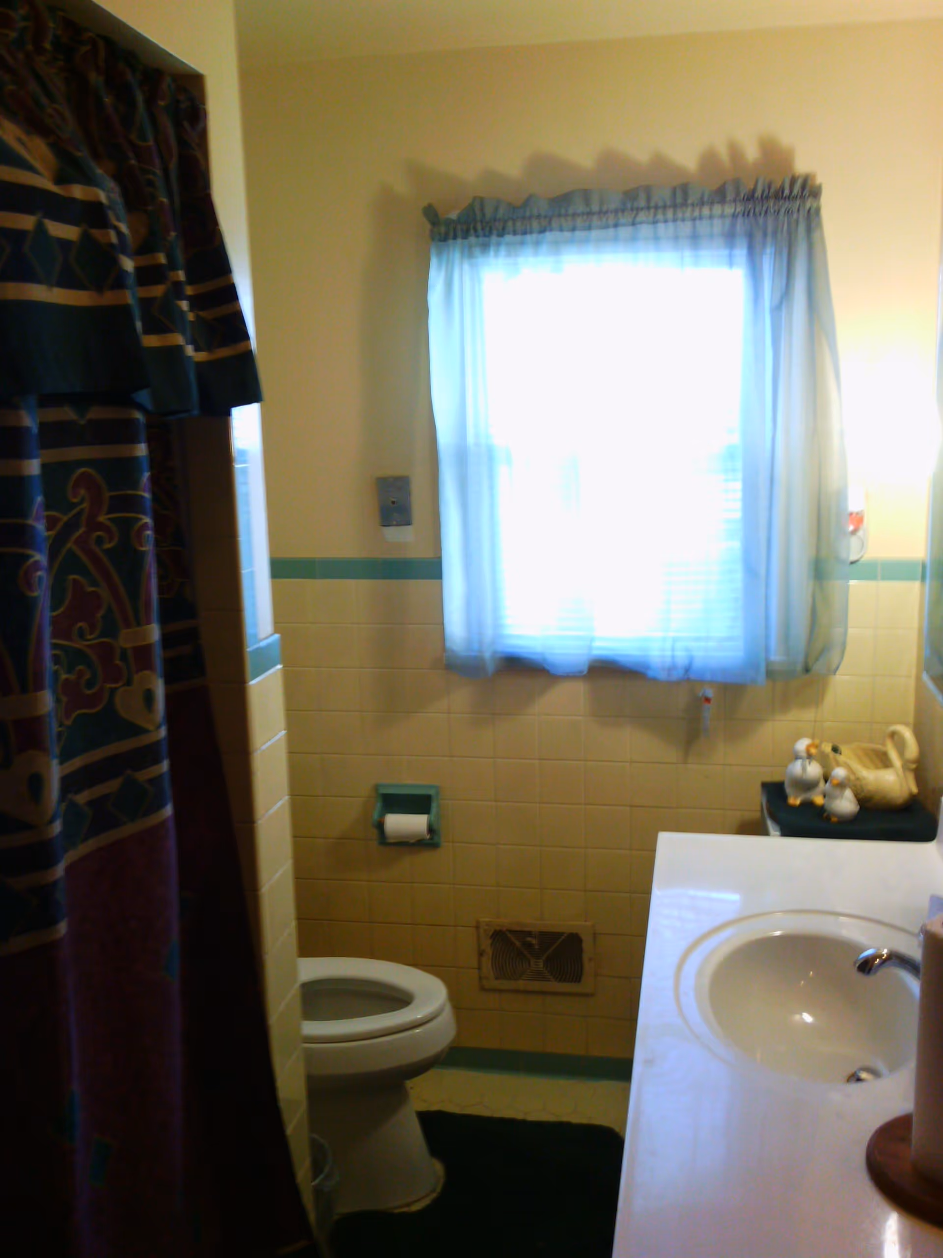A small bathroom with a toilet, a sink with a countertop, a window with sheer curtains, and a shower curtain with a colorful pattern. The walls are tiled halfway up in a light yellow color with a blue trim. There are decorative duck figurines on the countertop next to the sink.