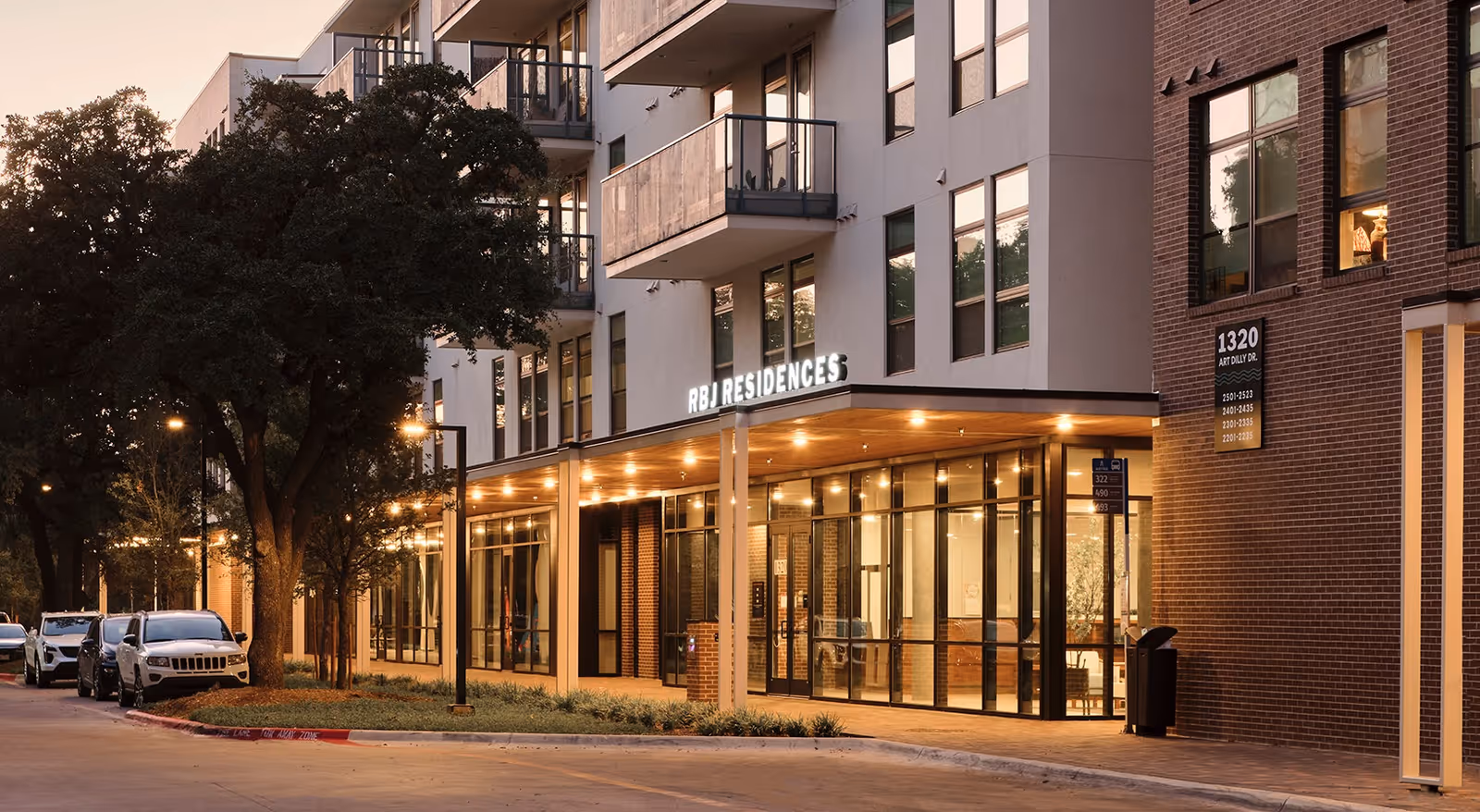 A modern senior housing building with a glass-front entrance and warm string lights under a canopy at dusk.
