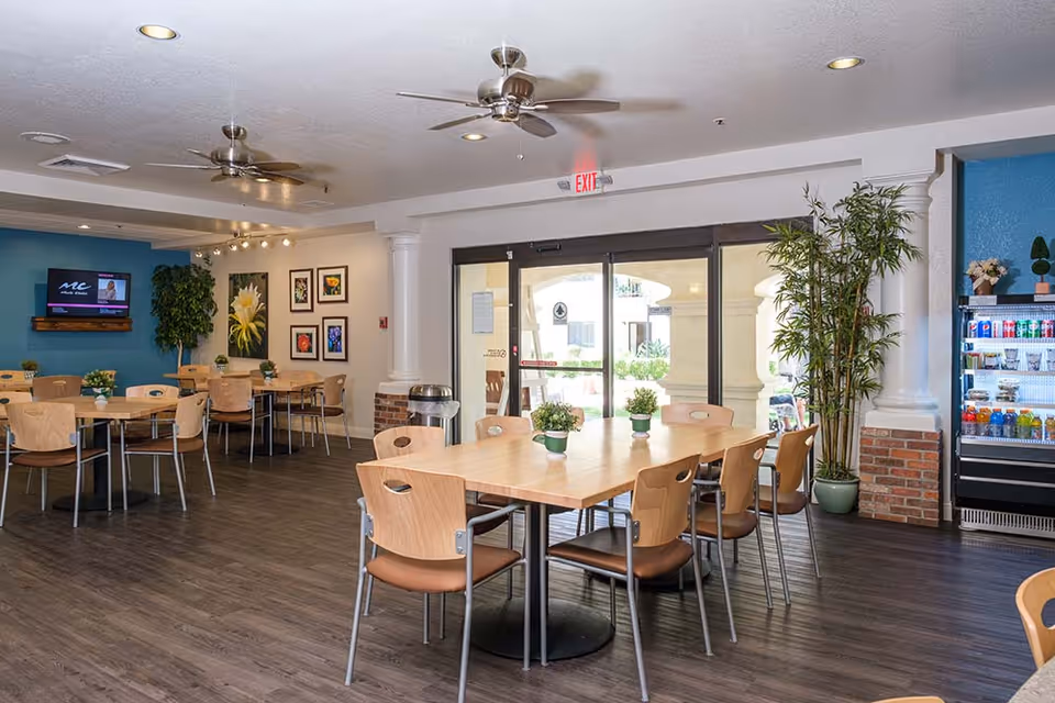 A bright and clean dining area with several wooden tables and chairs arranged neatly. There are potted plants on the tables and a large potted plant near the glass doors leading outside. The walls are decorated with framed floral artwork, and a television is mounted on a blue accent wall. A refrigerated display case with beverages is visible on the right side. Ceiling fans and recessed lighting illuminate the space.