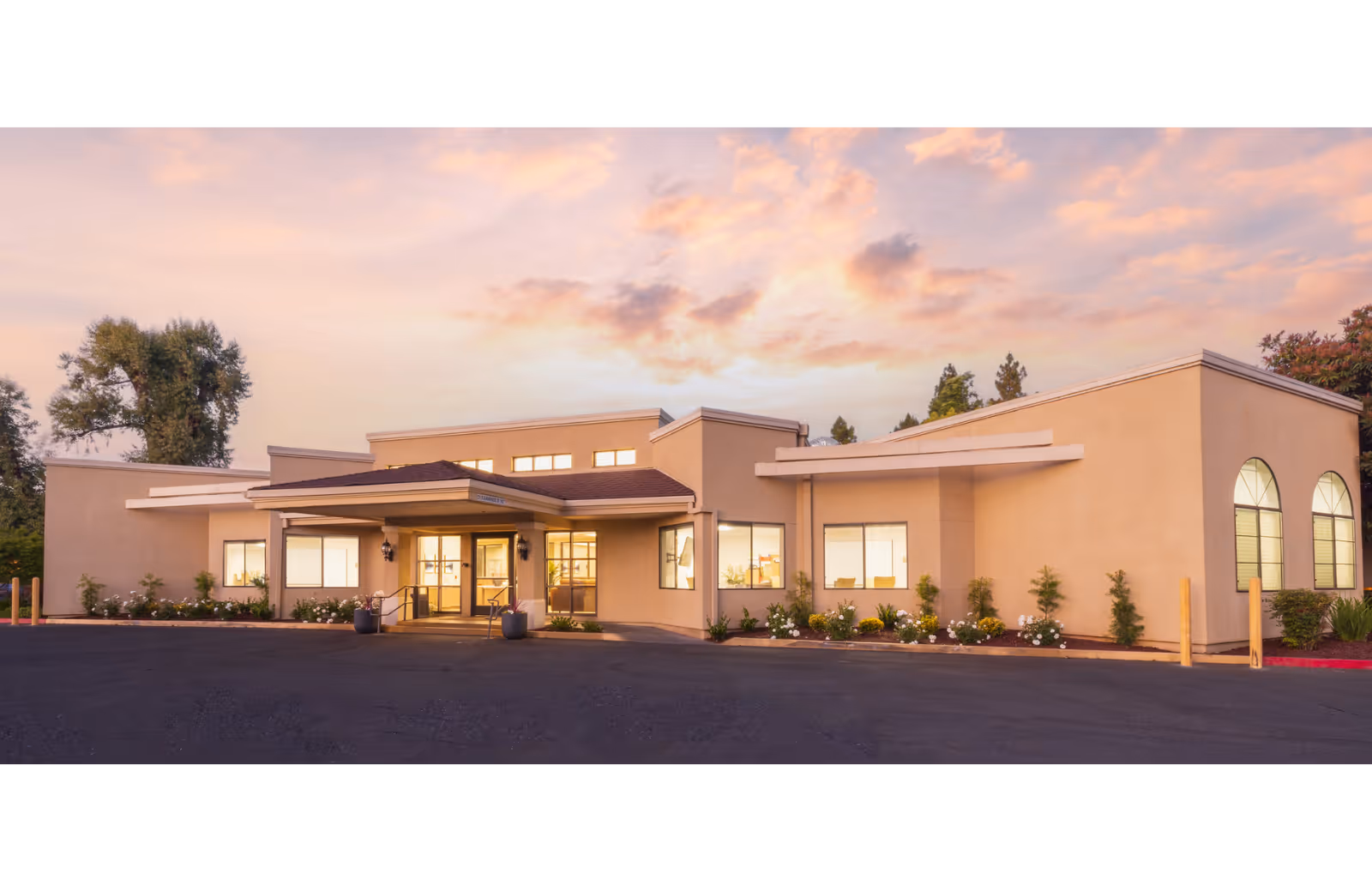 Front entrance of a single-story beige building with lit windows, landscaping, and a paved driveway under a colorful sunset sky.