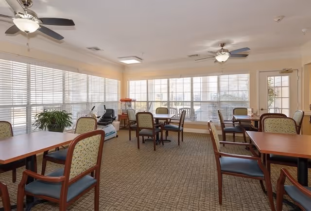A bright, spacious room with large windows covered by white blinds, multiple wooden tables with cushioned chairs arranged around them, two ceiling fans with lights, a potted plant, and an exercise machine near the windows.