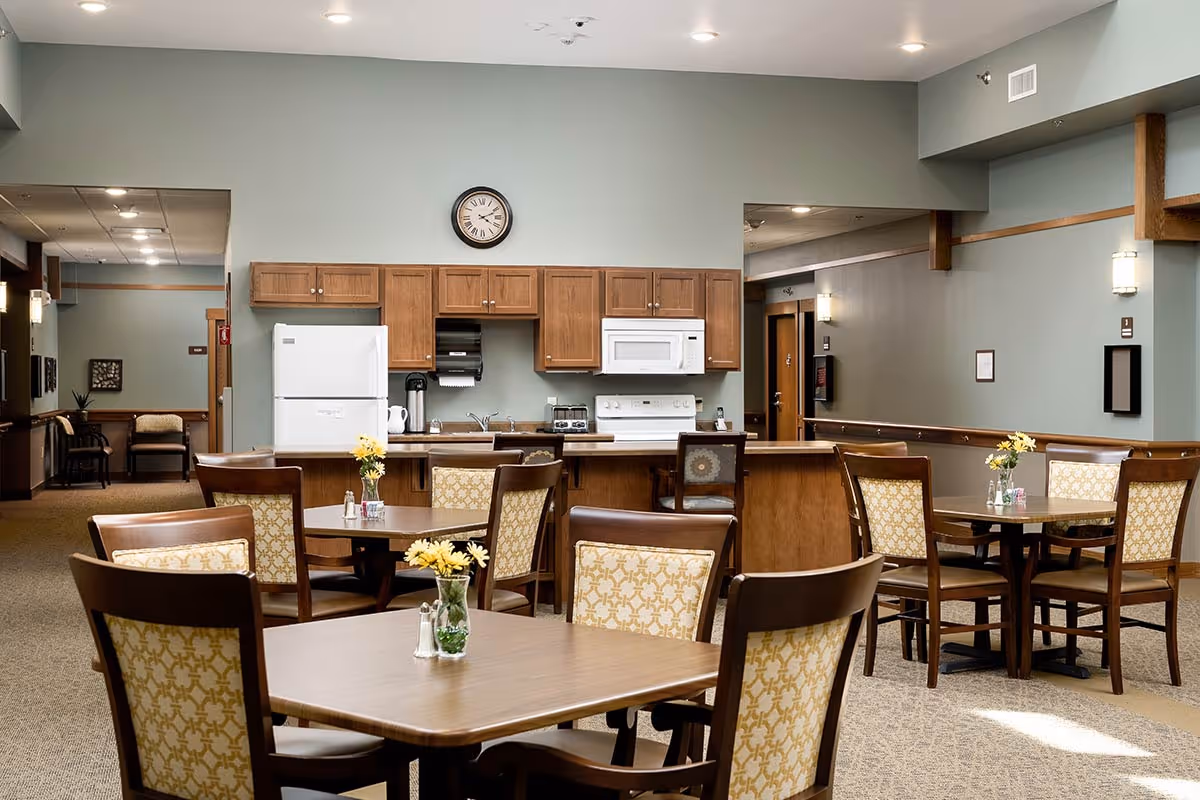 A senior living facility dining area with several wooden tables and chairs featuring patterned upholstery. Each table has a small vase with yellow flowers and salt and pepper shakers. In the background, there is a kitchen area with wooden cabinets, a white refrigerator, microwave, stove, and a clock on the wall above the cabinets. The walls are painted a soft green color, and the room is well-lit with ceiling lights.