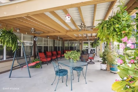 Covered outdoor patio area with hanging plants, ceiling fans, a swing, red cushioned chairs lined up against the wall, and a metal table with chairs in the center. There are various potted plants and flowers around the space.