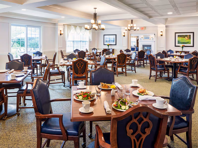 A spacious dining room with multiple wooden tables and chairs arranged neatly. Each table is set with plates of food, cups, glasses, and napkins. The room has large windows allowing natural light, elegant chandeliers, framed artwork on the walls, and a fireplace in the background.