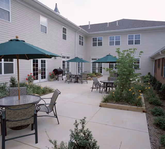Outdoor courtyard area of a senior living facility with several round glass tables and chairs, each shaded by green umbrellas. The courtyard is surrounded by a two-story beige building with multiple windows and doors. There are landscaped garden beds with various plants and flowers along the edges of the concrete patio.