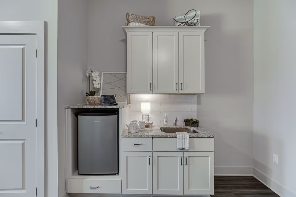 A small kitchenette area with white cabinets, a granite countertop, a mini refrigerator, a small sink, a soap dispenser, a small lamp, and decorative items including a basket, a plant, and a framed picture.