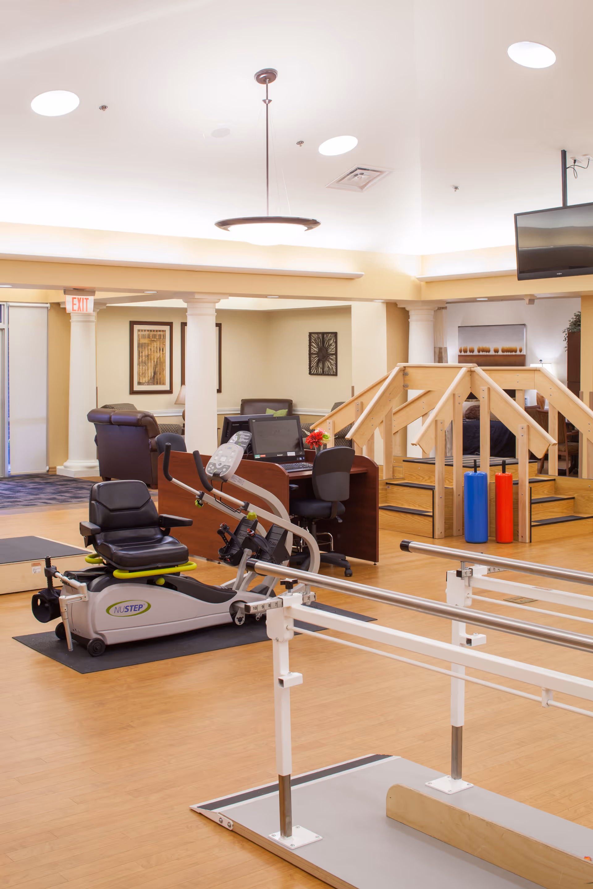 Interior view of a rehabilitation or physical therapy room in a senior living facility. The room features exercise equipment including a NuStep recumbent cross trainer, parallel bars for walking practice, and a wooden ramp with handrails. There is a desk with a computer and office chair in the background, along with comfortable seating and framed artwork on the walls. The space is well-lit with ceiling lights and has a warm, inviting atmosphere.