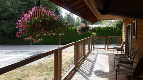 A covered wooden porch with hanging flower baskets, American flags, and chairs overlooking a driveway and hedge.
