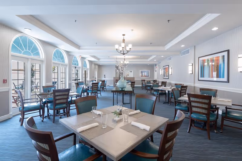 A spacious and well-lit dining room in a senior living facility with multiple tables and chairs arranged neatly. The room features large arched windows letting in natural light, blue cushioned chairs, a chandelier, framed artwork on the walls, and a central table with decorative vases and flowers.