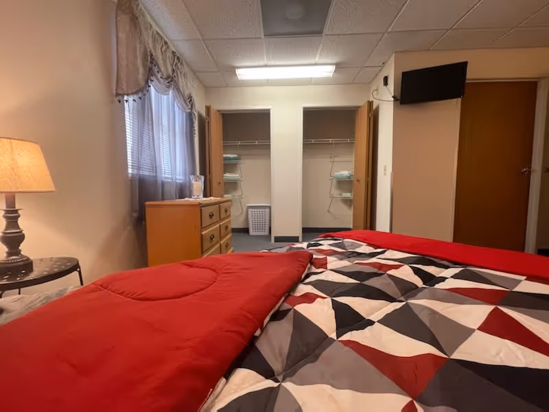 A bedroom with a bed covered in a geometric patterned quilt in red, black, white, and gray. There is a wooden dresser with folded towels on top, a laundry basket inside an open closet with shelves holding more towels, a small round table with a lamp, a window with curtains, and a wall-mounted TV above a closed door.
