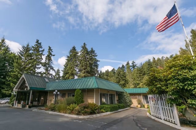 Exterior view of Bainbridge Island Health & Rehab Center, a single-story building with a green metal roof surrounded by trees and landscaping. An American flag is flying on a flagpole near the entrance.