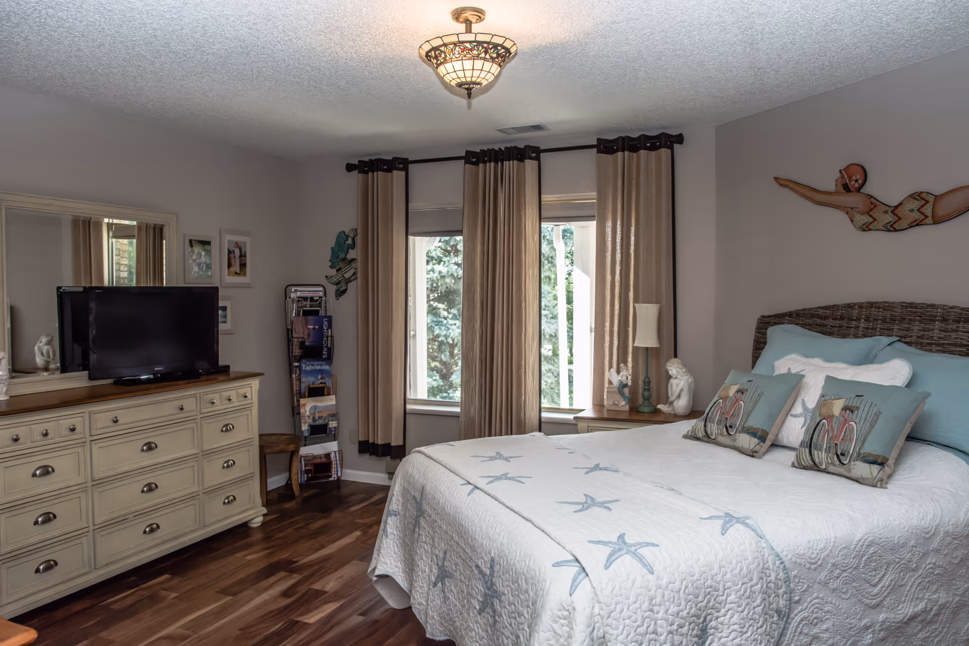 A cozy bedroom with a large bed covered in a white quilt with blue starfish patterns and decorative pillows featuring bicycles. The room has wooden flooring, beige walls, and a window with beige curtains. There is a dresser with a TV on top and a large mirror above it. A small side table with a lamp and decorative figurines is next to the bed. A wall decoration of a swimmer is mounted above the bed.