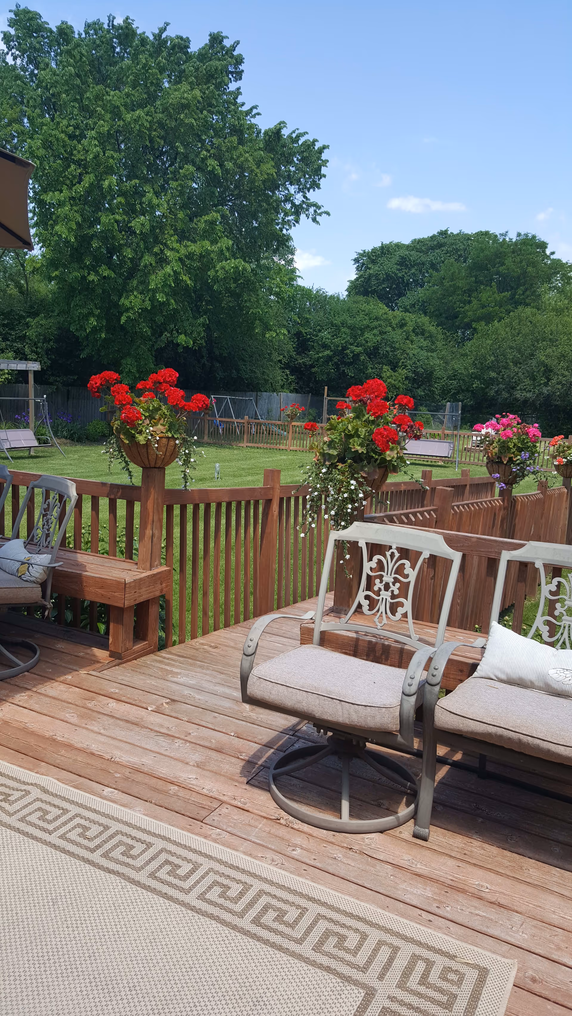 Wooden deck with cushioned metal chairs and a beige patterned outdoor rug. The deck railing is decorated with hanging flower baskets containing red and pink flowers. Beyond the deck is a green lawn with a wooden fence, trees, and a swing set under a clear blue sky.