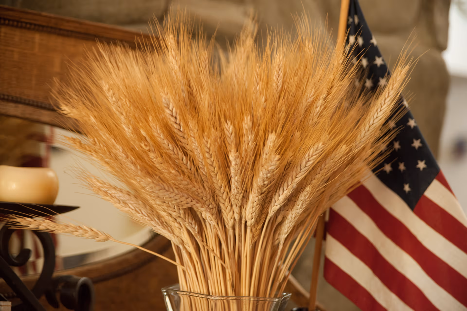 A close-up view of a glass vase filled with dried wheat stalks, positioned next to a small American flag. In the background, there is a wooden framed mirror and a candle on a decorative holder.