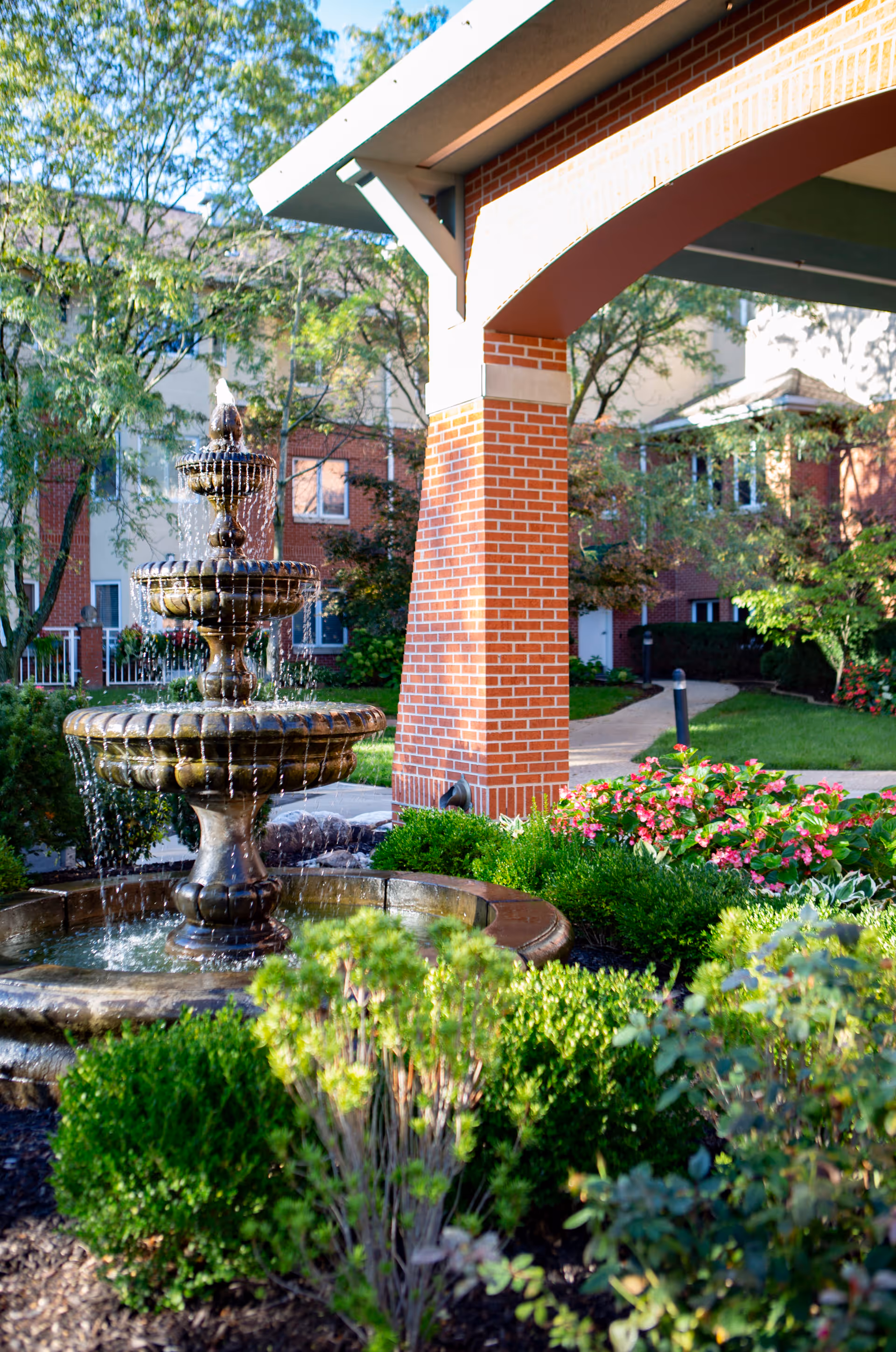A three-tiered water fountain surrounded by green bushes and flowering plants in a garden area. In the background, there is a brick pillar supporting a covered walkway, with a building and trees visible further behind.