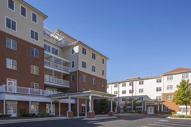 Exterior view of a multi-story senior living facility building with a covered entrance, multiple windows, and a clear blue sky.
