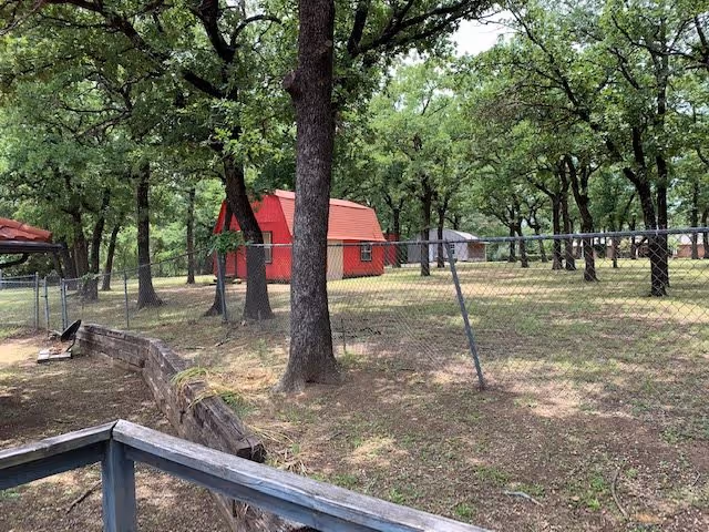 Outdoor fenced area with several trees and a small red barn-like structure in the background. The ground is mostly dirt with some grass patches, and there is a wooden retaining wall and a metal railing in the foreground.