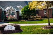 Exterior view of a brick building with a well-maintained lawn, shrubs, and a tree. A large rock in the foreground has a sign that reads 'The Woodlands Independent Suites 401-408'.