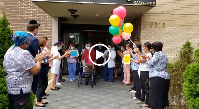 Staff and visitors line the entrance of Gold Crest Care Center applauding as a person using a walker exits holding colorful balloons.