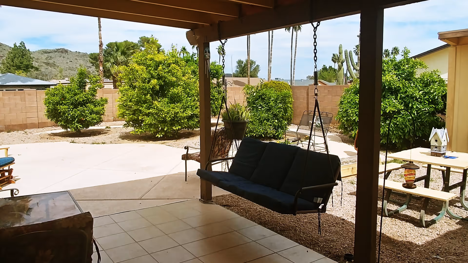 Covered backyard patio with a hanging porch swing, tiled flooring, picnic table, and shrubs against a block wall.