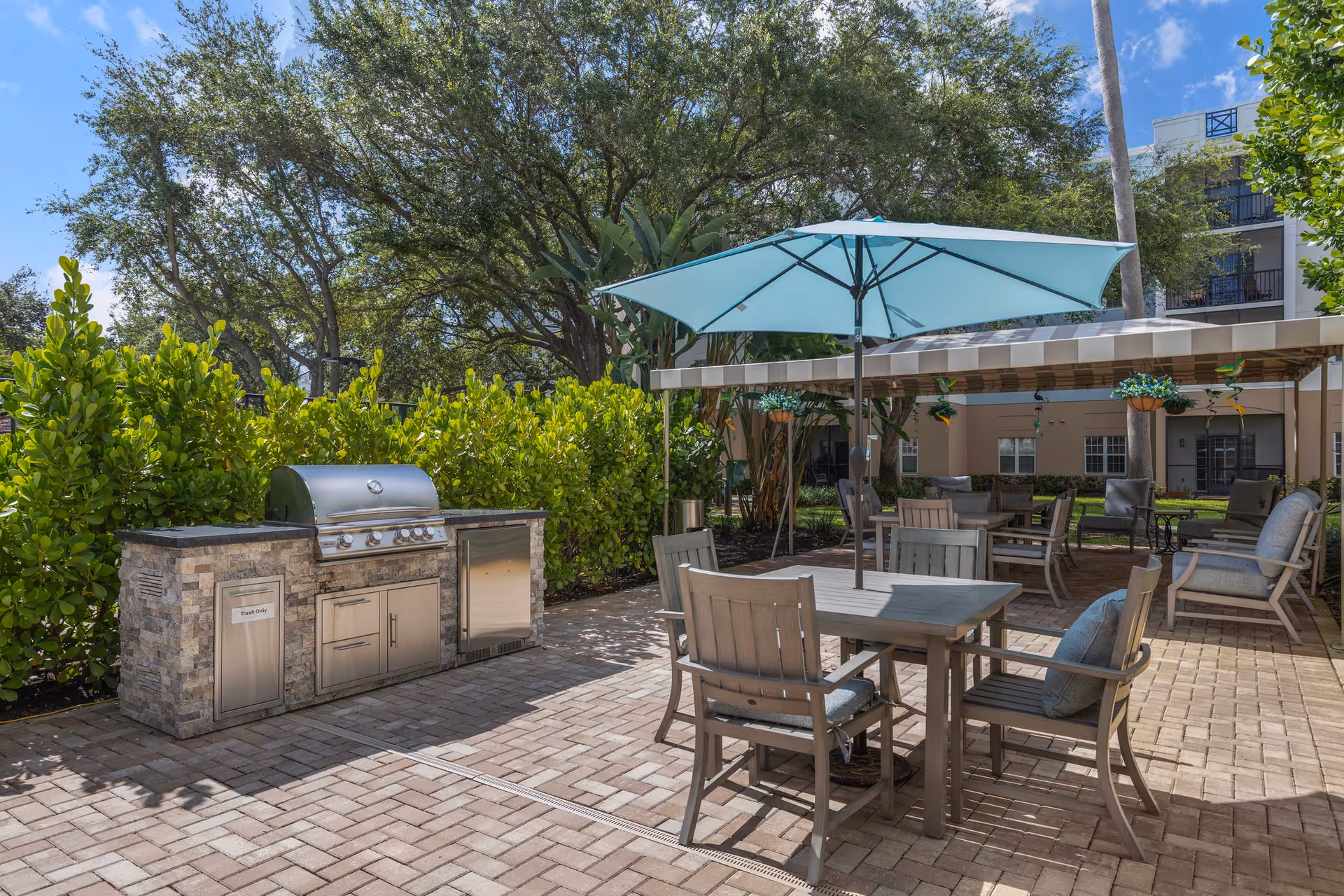 Outdoor patio area with a built-in stone grill and stainless steel appliances, surrounded by green bushes and trees. There are several wooden tables and chairs with cushions, one table shaded by a large blue umbrella. In the background, there is a covered seating area with more chairs and hanging plants, and a building is visible behind the patio.