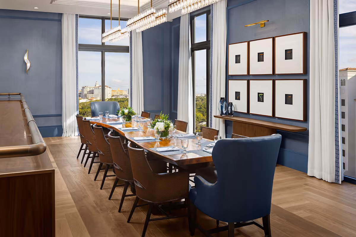 Elegant dining room with a long wooden table set for a meal, surrounded by wicker chairs and two blue upholstered armchairs at the ends. The room features large windows with white curtains, blue walls, modern light fixtures, framed artwork, and a view of buildings and greenery outside.