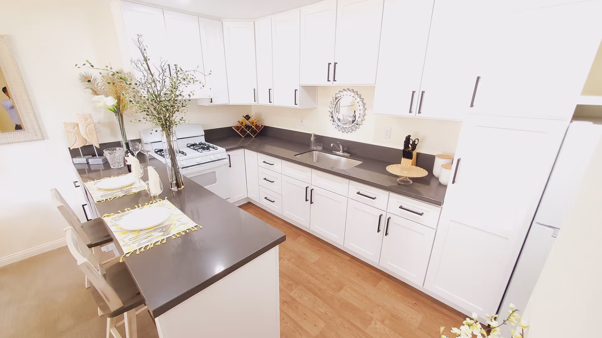 Bright and modern kitchen with white cabinets, gray countertops, a gas stove, and a sink. There is a breakfast bar with two chairs set with plates and napkins. Decorative plants and a round mirror are on the wall above the sink.